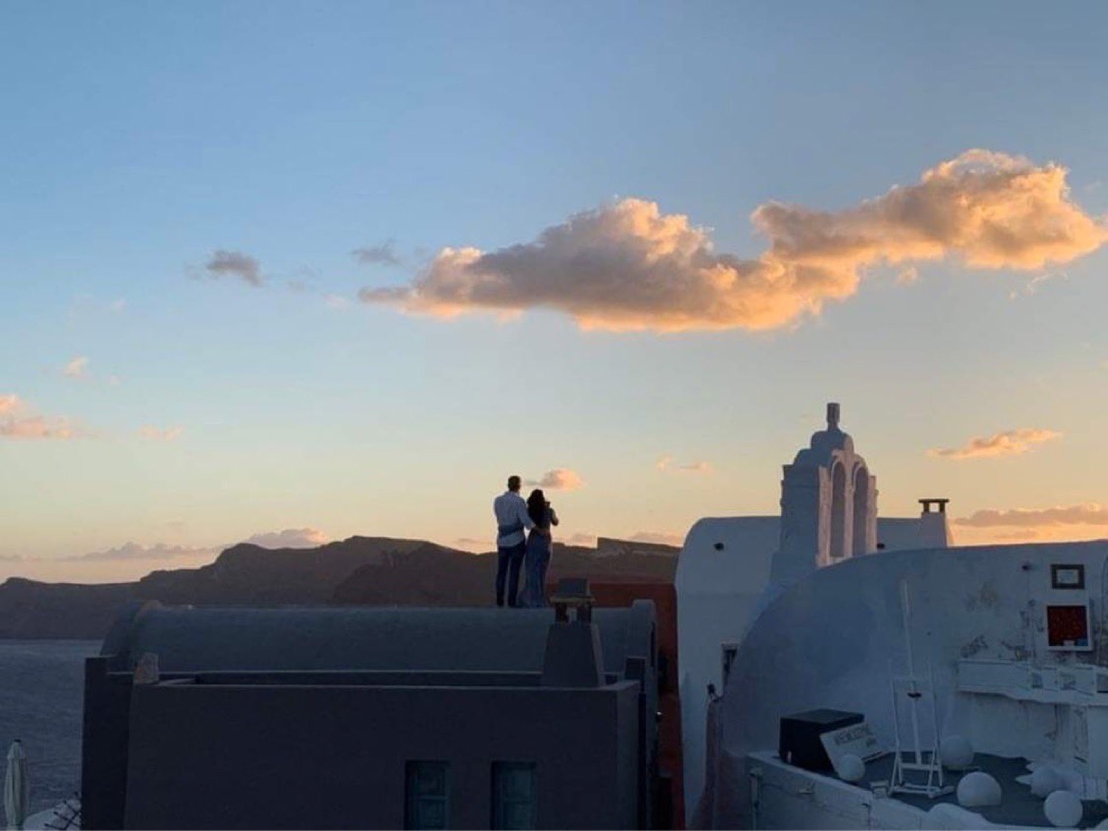 A couple standing on a rooftop at sunset, with whitewashed buildings to the right and the sea and hills behind
