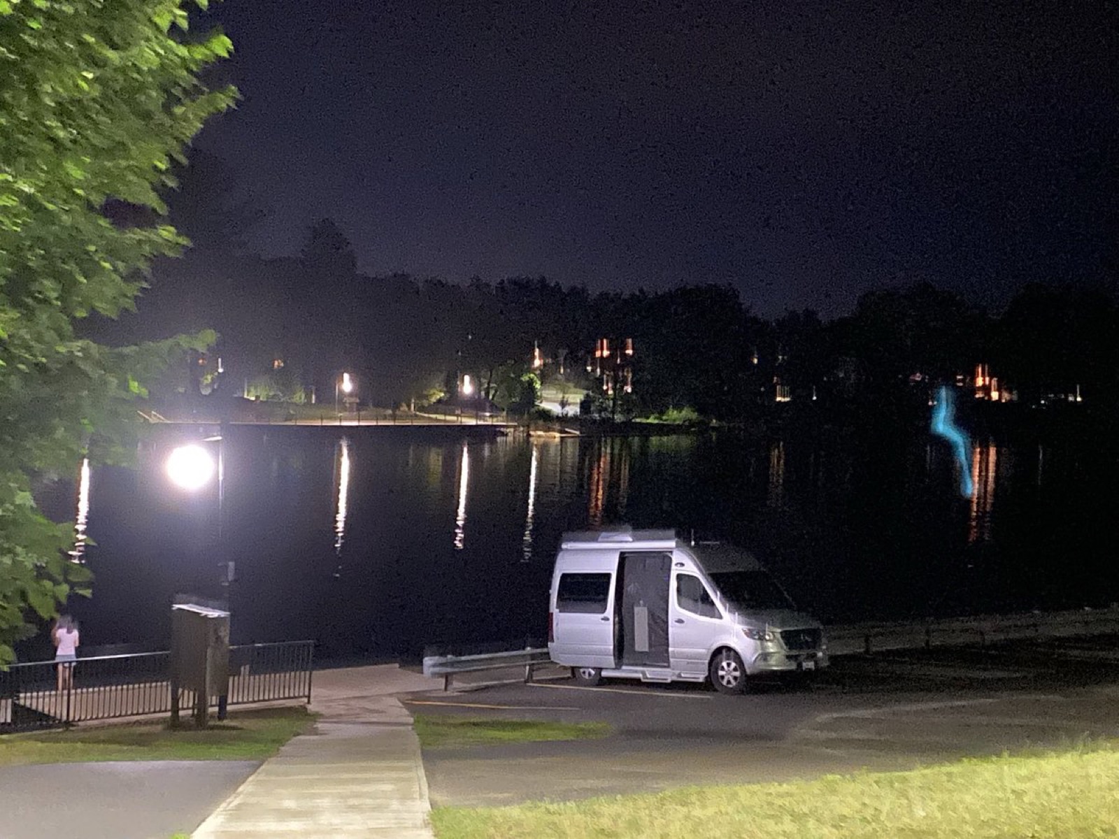 夜晚的湖边停车场，一辆银色的 B 型房车停在路灯下，对岸灯火倒映在水里