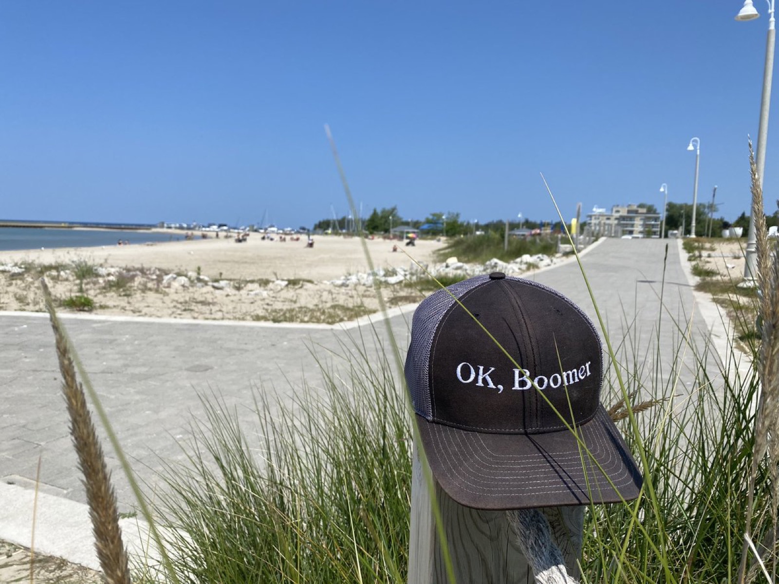 A dark brown trucker cap embroidered OK, Boomer, hung on a wooden post among beach grass, with a sandy beach and boardwalk behind