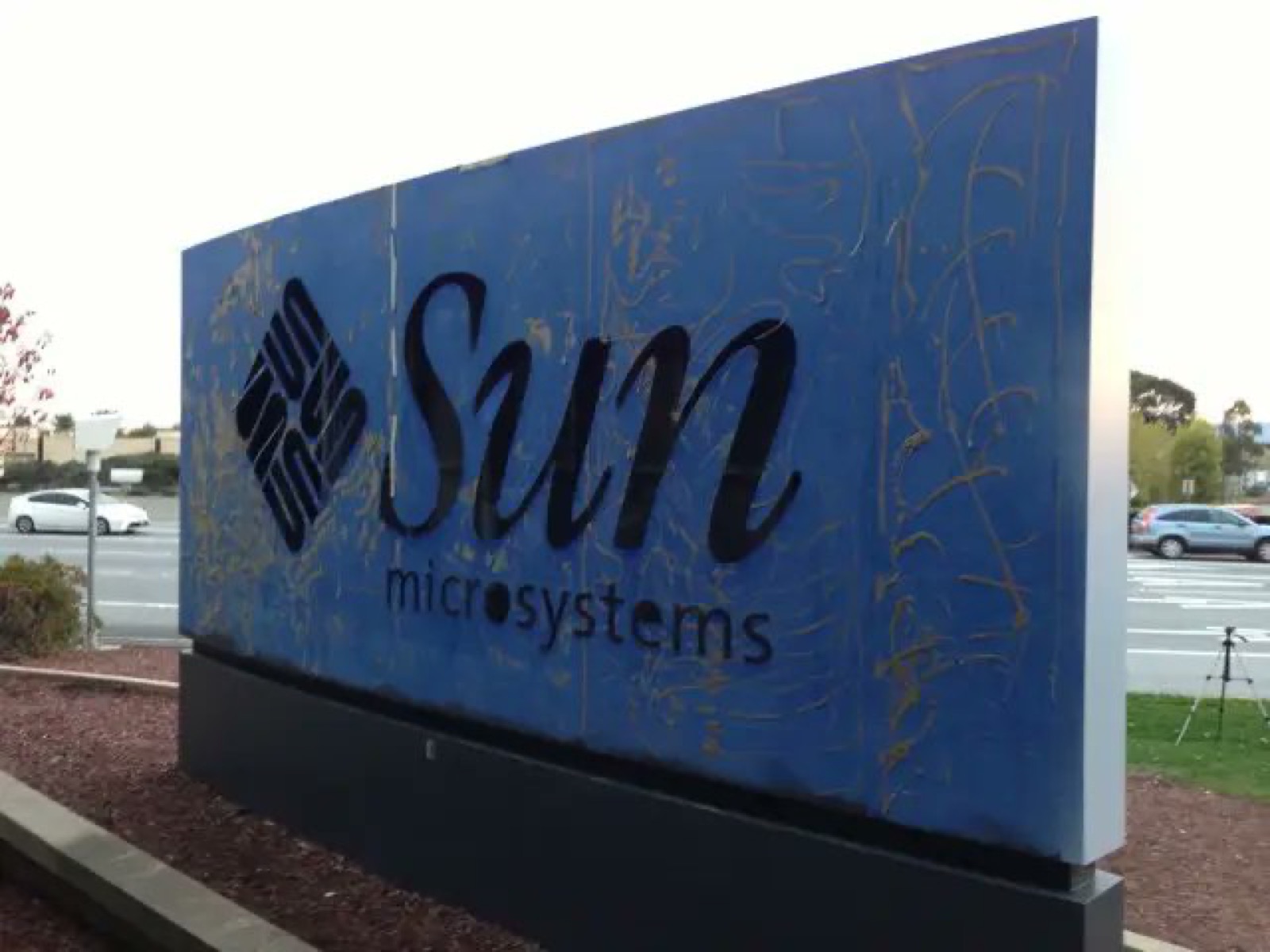A large blue roadside sign with the Sun Microsystems logo, its paint scratched and weathered
