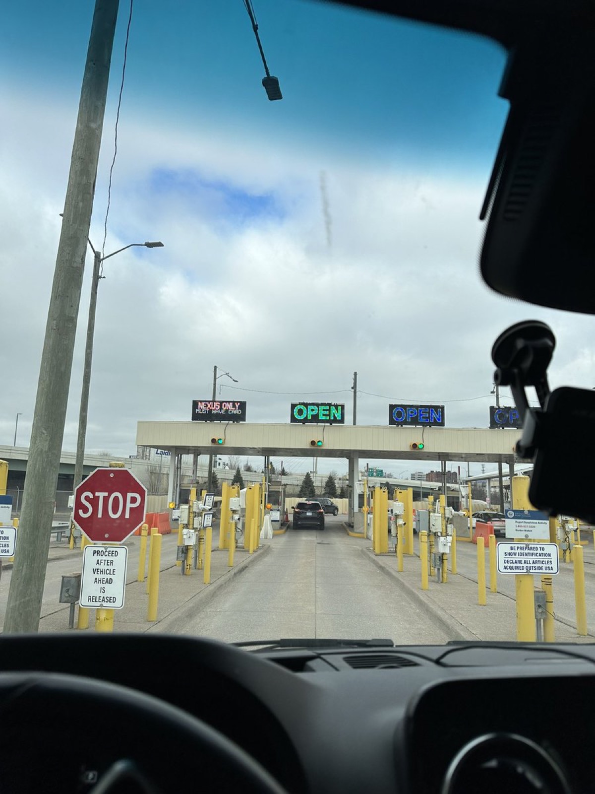 View through a camper van windscreen of a quiet border crossing: a stop sign, yellow bollards and booths marked OPEN under a cloudy sky