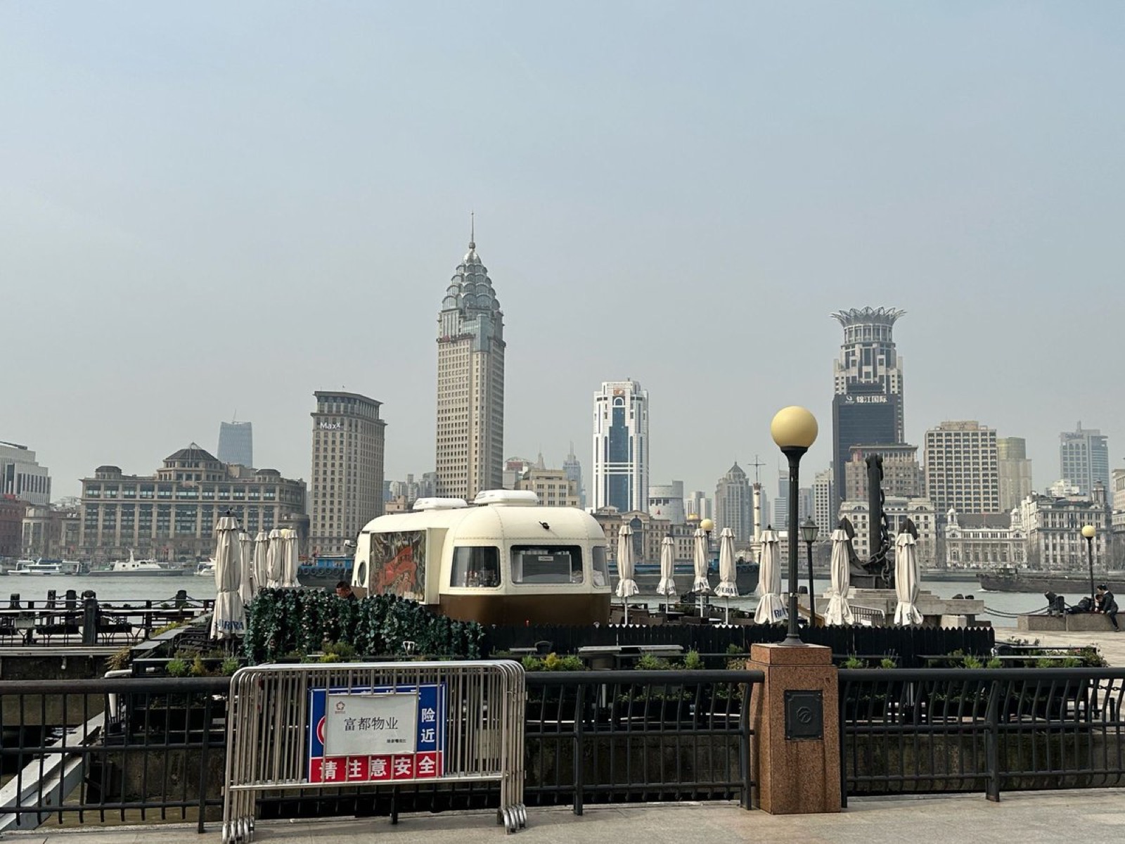 A rounded cream-and-brown vintage-style trailer parked on a riverside terrace beside closed white umbrellas, with the towers of the Shanghai Bund across the river behind it
