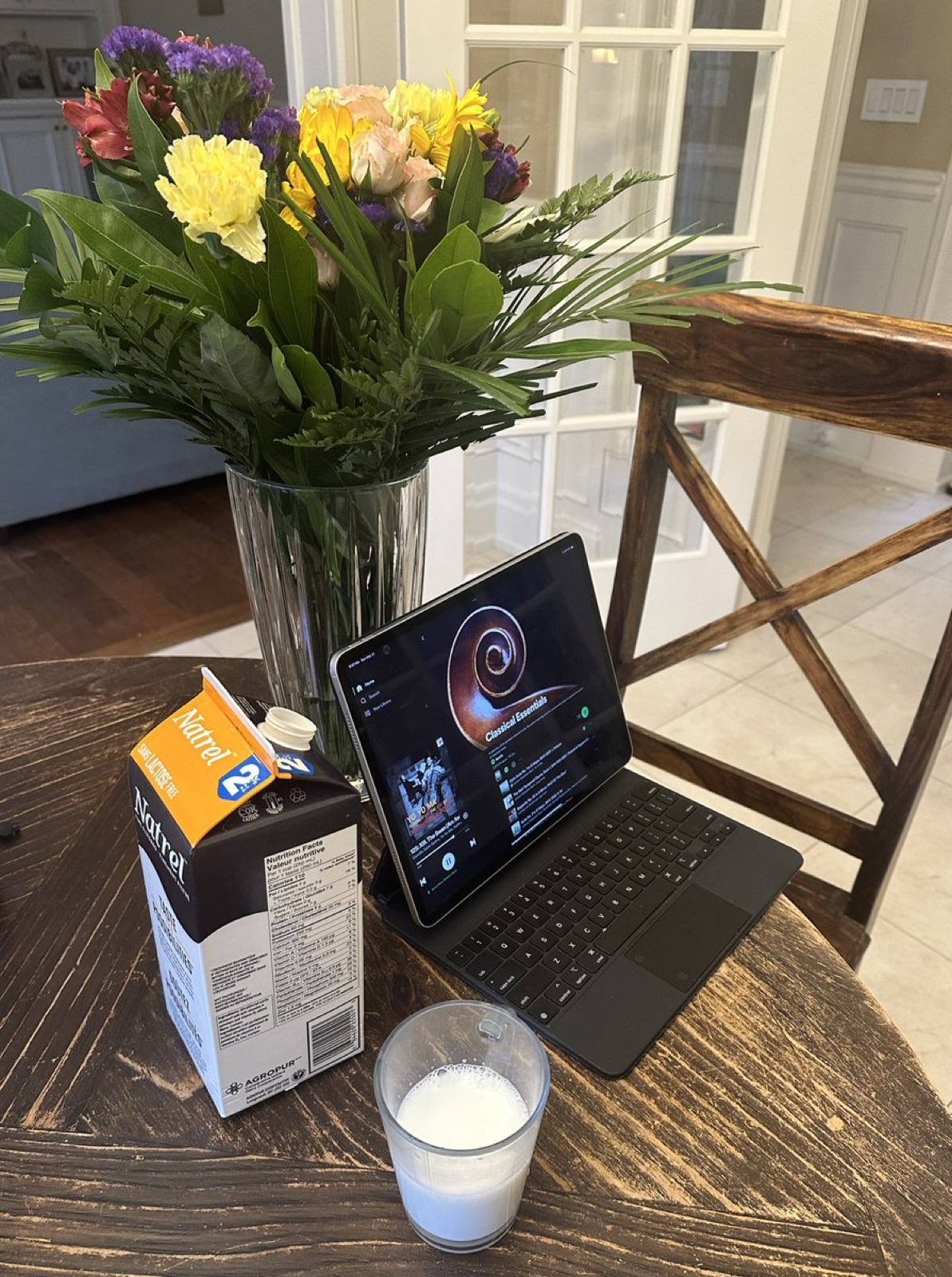 A carton and a glass of milk, a vase of mixed flowers and an iPad with a keyboard case on a wooden table